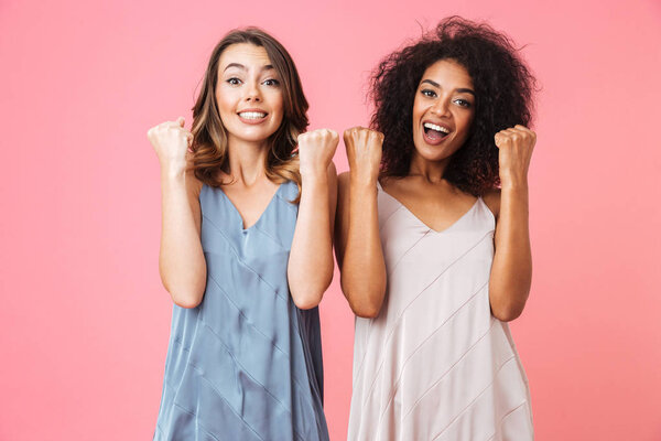 Two excited young girls dressed in summer clothes celebrating success isolated over pink background
