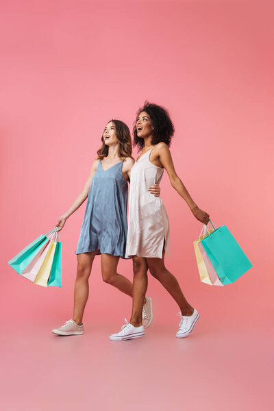 Full length portrait of two smiling young girls dressed in summer clothes holding shopping bags while walking isolated over pink background