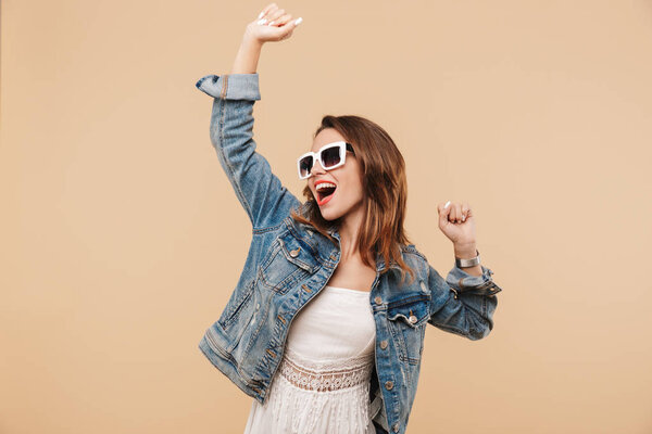 Portrait of a smiling young girl in summer clothes and sunglasses posing while standing with arms raised isolated over beige background
