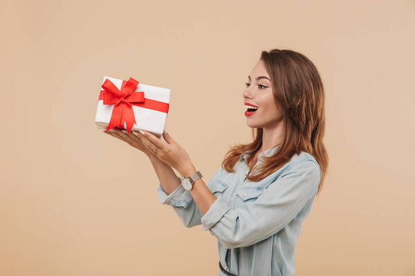 Portrait of an excited young girl looking at present box isolated over beige background