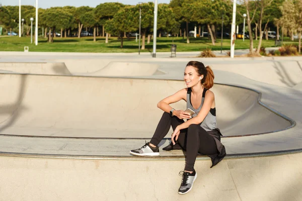 Image of young happy sports lady sitting outdoors in park using mobile phone looking aside listening music.