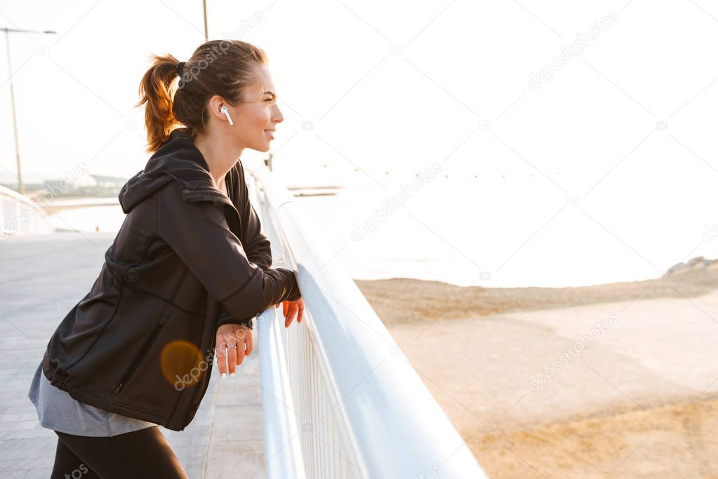Image of happy young sports woman standing outdoors on the street listening music with earphones.