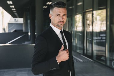 Yakışıklı bir genç işadamı portresi cam binanın önünde duran ve başparmak gösterilmesini takım elbise giymiş