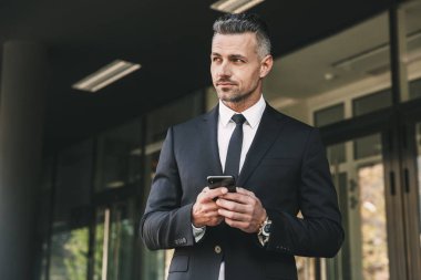 Yakışıklı bir genç işadamı portresi cam binanın önünde duran ve cep telefonu tutan takım elbise giymiş