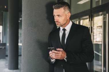 Kendine güvenen genç işadamı portresi cam binanın önünde duran ve cep telefonuyla takım elbise giymiş