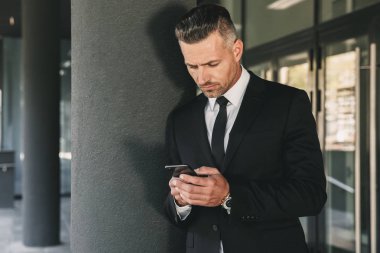 Konsantre bir genç işadamı portresi cam binanın önünde duran ve cep telefonuyla takım elbise giymiş