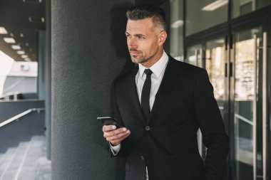 Başarılı bir genç iş adamı portresi cam binanın önünde duran ve cep telefonuyla takım elbise giymiş