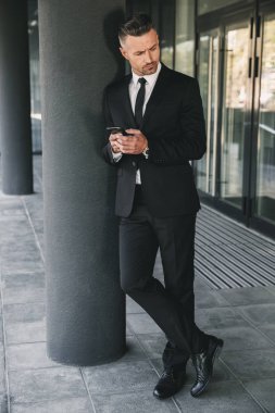 Konsantre bir işadamı portresi cam binanın önünde duran ve cep telefonuyla takım elbise giymiş