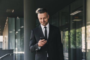 Büyüleyici bir genç işadamı portresi bina ve cep telefonuyla bir cam dışında yürüyüş takım elbise giymiş