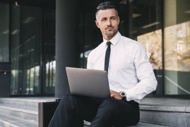 Resmi elbise bina ve dizüstü bilgisayar üzerinde çalışan bir cam dışında oturan başarılı bir genç iş adamı portresi giymiş