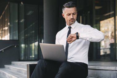 Dizüstü bilgisayar ile bina ve onun bilek bakarak bir cam dışında oturan resmi elbise giymiş odaklı bir genç işadamı portresi