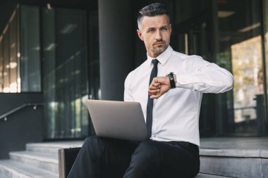 Resmi elbise dizüstü bilgisayar ile bina ve onun bilek bakarak bir cam dışında oturan kendine güvenen genç işadamı portresi giymiş