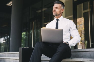 Resmi elbise bina ve dizüstü bilgisayar üzerinde çalışan bir cam dışarıda gülümseyen genç işadamı portresi giymiş