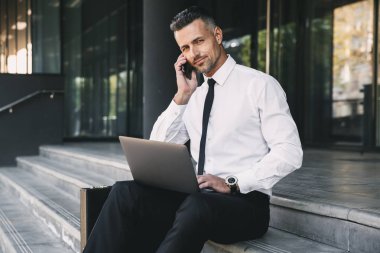 Resmi elbise bina ile dizüstü bilgisayar ve cep telefonu üzerinde konuşurken bir cam dışında oturan kendine güvenen genç işadamı portresi giymiş