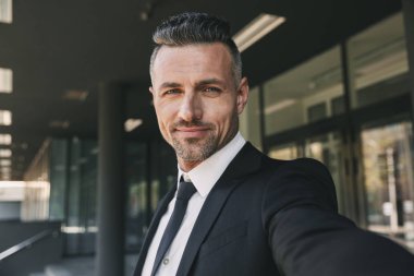 Gülümseyen genç işadamı portresi cam binanın önünde duran ve bir selfie alarak takım elbise giymiş