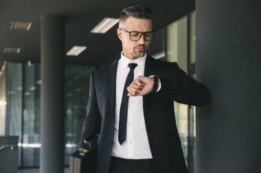 Başarılı bir genç iş adamı portresi cam binanın önünde duran ve onun bilek bakarak takım elbise giymiş