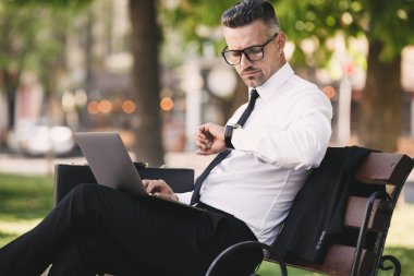 Yakışıklı bir genç işadamı portresi dışında bir bankta oturan ve dizüstü bilgisayar üzerinde çalışan resmi elbise giymiş
