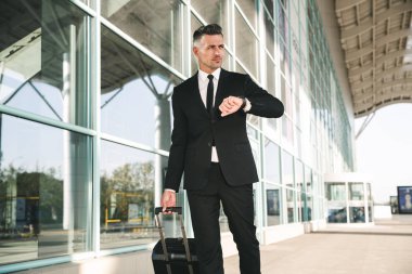 Rahat işadamı ile bir bavul Havaalanı terminal dışında yürüyüş ve kol seyir takım elbise giymiş