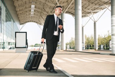 Yakışıklı işadamı ile bir bavul Havaalanı terminal dışında yürüyüş ve cep telefonu tutan takım elbise giymiş