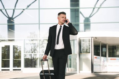 Yakışıklı işadamı ile bir bavul Havaalanı terminal dışında yürüyüş ve cep telefonu üzerinde konuşurken takım elbise giymiş