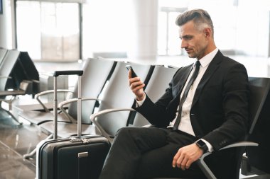 Bir bavulla Havaalanı lobisinde otururken cep telefonuyla yakışıklı olgun işadamı