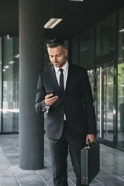 Portrait of a handsome young businessman dressed in suit with briefcase walking outside a glass building and using mobile phone