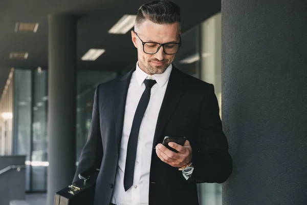 ブリーフケース ガラスの建物の外に立って 携帯電話を見ているとスーツに身を包んだハンサムな青年実業家の肖像画 — ストック写真