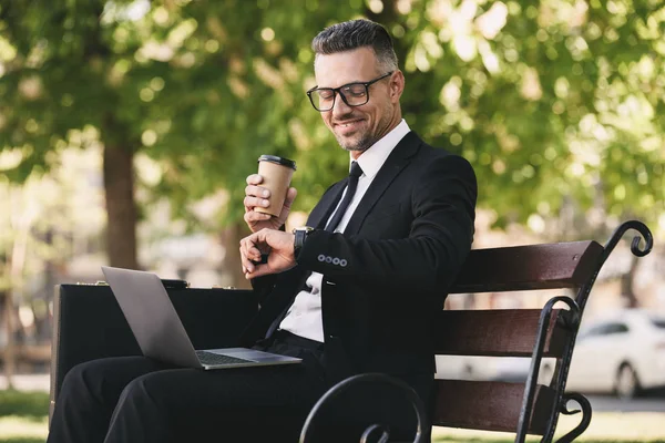 Portrait of a smiling businessman dressed in formal clothes sitting outside on a bench with laptop computer and drinking coffee