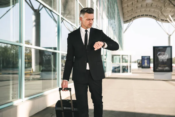Serious businessman dressed in suit walking with a suitcase outside airport terminal and looking at his wristwatch