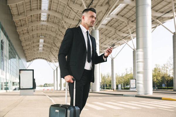Confident businessman dressed in suit walking with a suitcase outside airport terminal and holding mobile phone