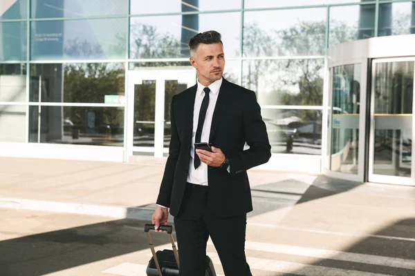 Handsome businessman dressed in suit walking with a suitcase outside airport terminal and holding mobile phone