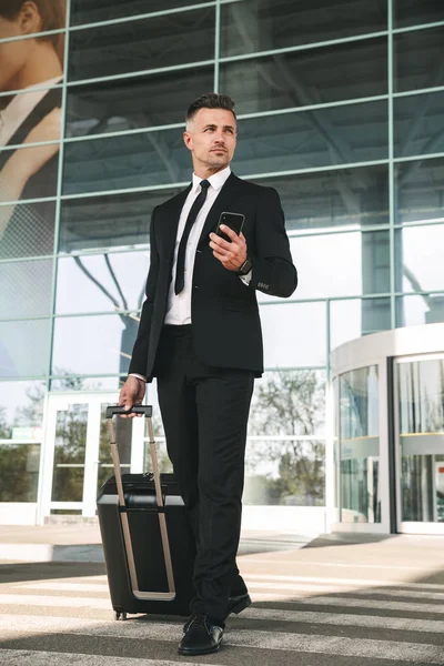 空港ターミナルの外のスーツケースを持って歩くと 携帯電話を保持しているスーツを着ている集中ビジネスマン — ストック写真