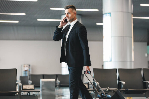 Attractive mature businessman talking on mobile phone while walking at the airport lobby with a suitcase