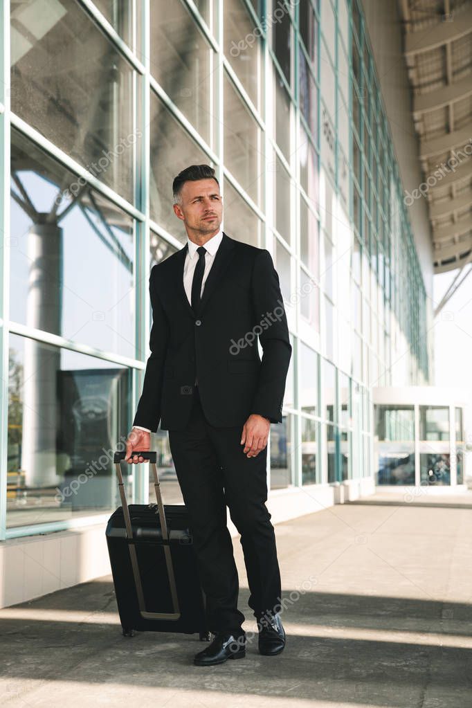 Charming businessman dressed in suit walking with a suitcase outside airport terminal