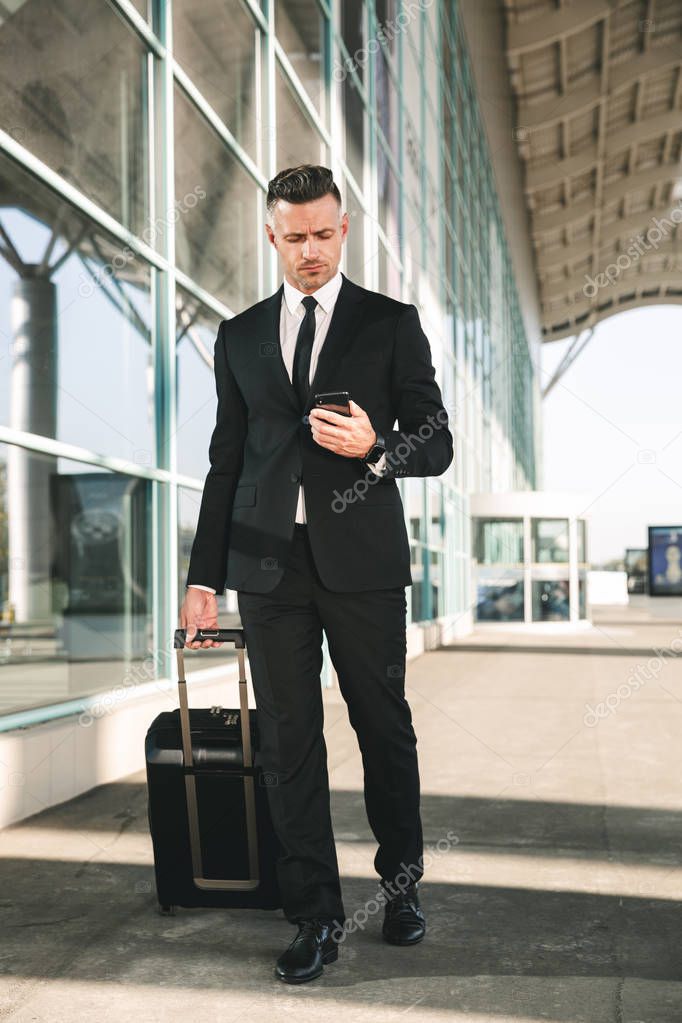 Confident businessman dressed in suit walking with a suitcase outside airport terminal and looking at mobile phone