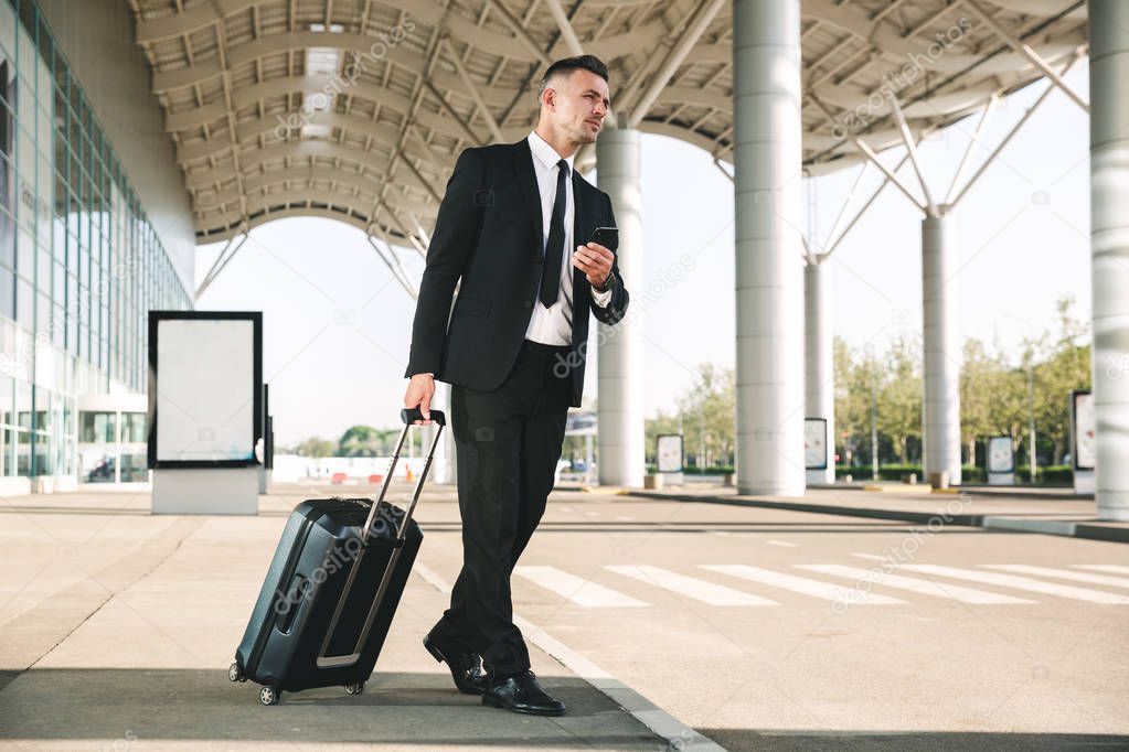 Handsome businessman dressed in suit walking with a suitcase outside airport terminal and holding mobile phone