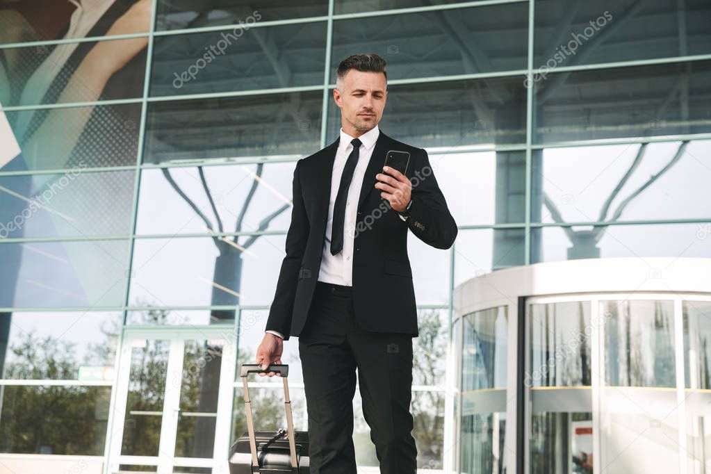 Serious businessman dressed in suit walking with a suitcase outside airport terminal and holding mobile phone