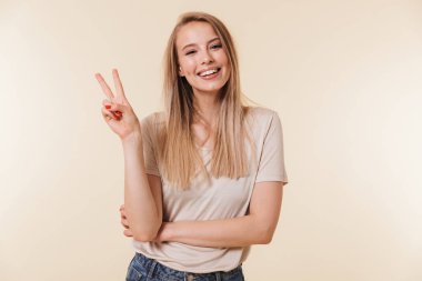 Casual giyim gülümseyen ve barış işareti Studio bej renkli arka plan üzerinde izole iki parmak ile gösterilen giyen neşeli beyaz kadın görüntüsünü