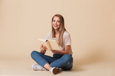 Bacaklar ile katta oturan rahat giysiler giyiyor neşeli gülümseyen kadın fotoğrafı geçti ve komik kitap okuma bej renkli arka plan üzerinde izole