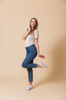 Tam uzunlukta portre çekici sarışın kadın 20'li yaşlarda giyiyor rahat giyim gülümseme ile kamera poz ve bej renkli arka plan üzerinde izole onun bacak kaldırma