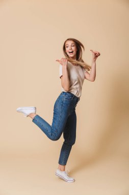 Tam boy görüntü çalıştıran ve bej renkli arka plan üzerinde izole boşaltmak, parmakları geriye doğru işaret memnun sarışın kadın 20'li yaşlarda giyiyor rahat giyim