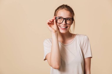 Görüntü güzel neşeli kadının temel giyen 20s Studio bej renkli arka plan üzerinde gözlük dokunmaktan ve bir kenara bakarken kameraya gülümseyen t-shirt izole