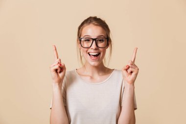 Temel t-shirt ve yukarı doğru Studio bej renkli arka plan üzerinde izole parmakları işaret ederken gülümseyen gözlük takıyor bağlı saçlı mutlu sarışın kadın 20'li yaşlarda görüntüsünü