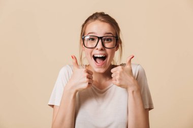 Photo of excited woman 20s with blonde tied hair wearing basic t-shirt and eyeglasses laughing while showing thumbs up isolated over beige background in studio clipart