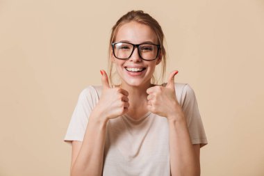 Photo of satisfied pleased woman 20s with blonde tied hair wearing basic t-shirt and eyeglasses smiling while showing thumbs up isolated over beige background in studio clipart