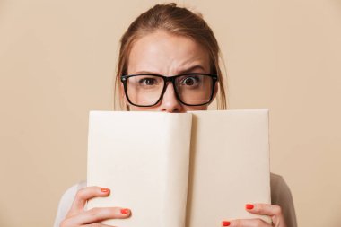 Kitap yüzüne tutarak ve bej renkli arka plan üzerinde izole kamera bakarak Eyewear bir genç kız portresi