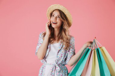 Yazlık elbise ve hasır şapka alışveriş torbaları üzerinde pembe arka plan izole tutarken cep telefonu üzerinde konuşurken mutlu bir genç kadın portresi