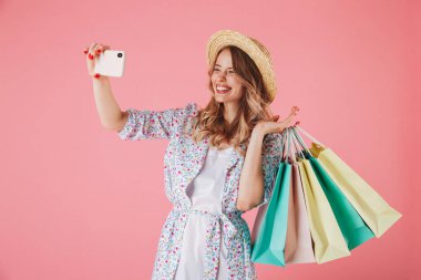 Yazlık elbise ve alışveriş torbaları üzerinde pembe arka plan izole tutarken bir selfie alarak hasır şapka neşeli bir genç kadın portresi