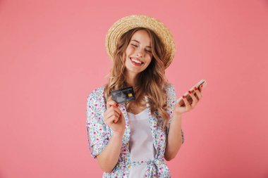 Yazlık elbise ve cep telefonu pembe arka plan izole kullanırken hasır şapka gösteren plastik kredi kartı neşeli bir genç kadının portresi
