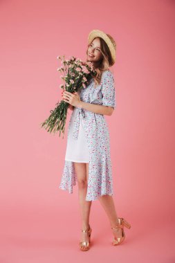 Yazlık elbise ve hasır şapka Karanfil Buketi pembe arka plan üzerinde izole tutarken poz güzel bir genç kadın portresi tam uzunlukta